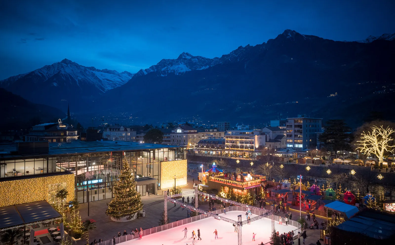 Panoramablick auf den Meraner Weihnachtsmarkt mit Eislaufbahn, Therme-Glasfassade und schneebedeckten Bergen zur blauen Stunde