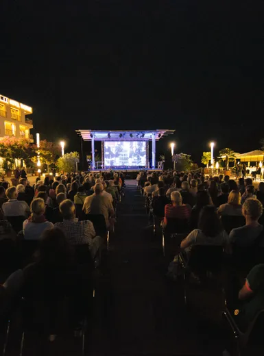 Abendliche Open-Air-Veranstaltung vor der Therme Meran mit Publikum auf Stühlen vor beleuchteter Bühne, umgeben von Palmen und warmem Gebäudelicht
